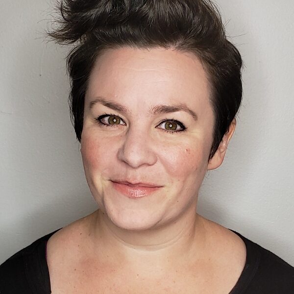 Writing coach Megan Cohen is a middle aged white cis woman with a swirling pouf of brown hair on top of her head that looks like a croissant. She smiles at the camera with a closed mouth.